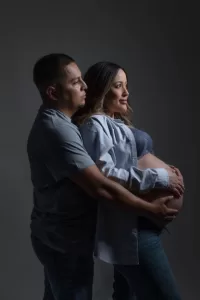 Professional Photographer A man stands behind a pregnant woman, gently holding her belly. Both wear casual clothes and look thoughtfully into the distance, as if posing for intimate headshots against a dark, neutral background. www.maranonimagery.com