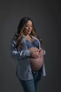 Professional Photographer A pregnant woman wearing a blue bra, unbuttoned striped shirt, and jeans gently cradles her bare belly with both hands, smiling softly against a plain gray background—perfect for maternity portraits. www.maranonimagery.com