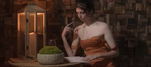 Professional Photographer A woman in a strapless, copper dress sits at a wooden table, reading a book. A decorative basket of moss and a glass of wine are on the table—an elegant scene reminiscent of fine wedding photography, with a lantern glowing warmly behind her. www.maranonimagery.com