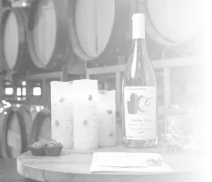 Professional Photographer A wine bottle, chocolates, birch-decorated candles, glasses, and a card are arranged on a wooden table in front of stacked wine barrels—perfect for wedding photography. The image features a soft white gradient on the right side. www.maranonimagery.com