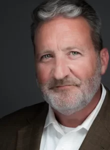 Professional Photographer A middle-aged man with short gray hair and a neatly trimmed beard, wearing a white shirt and brown blazer, poses for professional headshots against a dark gray background with a slight smile. www.maranonimagery.com