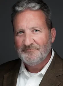 Professional Photographer A middle-aged man with gray hair and a short beard, wearing a brown blazer and white shirt, poses for a professional portrait against a dark gray background, looking directly at the camera with a slight smile. www.maranonimagery.com