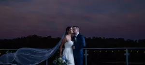 Professional Photographer A bride in a white gown and veil stands with a groom in a blue suit on an outdoor balcony at dusk, captured in elegant wedding photography. The bride holds a bouquet of blue and white flowers against a purple-blue sky. www.maranonimagery.com