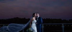 Professional Photographer A bride and groom stand close together on an outdoor balcony at dusk, her long veil flowing in the wind and the sky painted in purple and blue—a perfect moment of wedding photography, with a bouquet of blue and white flowers. www.maranonimagery.com