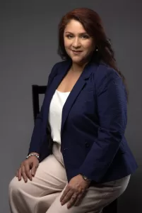Professional Photographer A woman with long dark hair sits on a chair, wearing a navy blazer, white blouse, and light-colored pants, posing confidently for a headshot against a plain dark background. www.maranonimagery.com