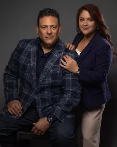 Professional Photographer A man in a blue plaid blazer and dark pants sits on a stool, while a woman in a navy blazer and light pants stands beside him with her hand on his shoulder—an elegant portrait captured by a talented photographer against a dark background. www.maranonimagery.com