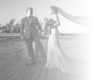 Professional Photographer A bride in a long gown and veil walks with a man in a suit on a wooden deck near water, holding a bouquet. Captured by a skilled photographer, the scene is partly faded with a bright, soft white overlay on the right side. www.maranonimagery.com