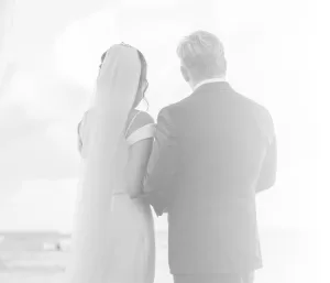 Professional Photographer A bride in a wedding dress and veil stands beside a man in a suit, seen from behind, as a wedding photographer captures dreamy portraits overlooking the softly lit beach or ocean. www.maranonimagery.com