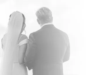 Professional Photographer A bride and groom stand side by side, seen from behind. Perfect for wedding photography, the bride’s veil and white dress with thin straps complement the groom’s suit against a softly lit, bright, airy background. www.maranonimagery.com