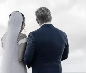 Professional Photographer A bride in a white dress and veil stands beside a groom in a dark suit, both facing away from the camera against a cloudy sky—capturing a timeless moment in wedding photography. www.maranonimagery.com