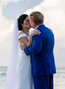 Professional Photographer A bride and groom, dressed in a white gown and blue suit, share a kiss outdoors by the sea under a bright sky, embracing each other on their wedding day—an unforgettable portrait capturing pure love. www.maranonimagery.com