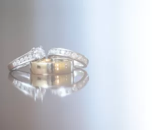 Professional Photographer Three rings rest on a reflective surface: one gold wedding band in front and two silver rings with diamonds behind it, beautifully styled to capture the elegance often seen in wedding photography against a softly lit gradient white background. www.maranonimagery.com