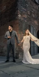 Professional Photographer A bride in a lace wedding dress and a groom in a gray suit hold hands near a brick building with arched doors, both gazing in different directions as their photographer perfectly captures the moment. www.maranonimagery.com