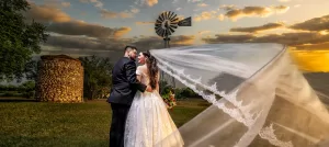 Professional Photographer A bride and groom embrace outdoors at sunset, their long veil flowing in the wind. A photographer captures this romantic moment near a windmill and stone building under a partly cloudy sky—beautiful memories in portraits. www.maranonimagery.com