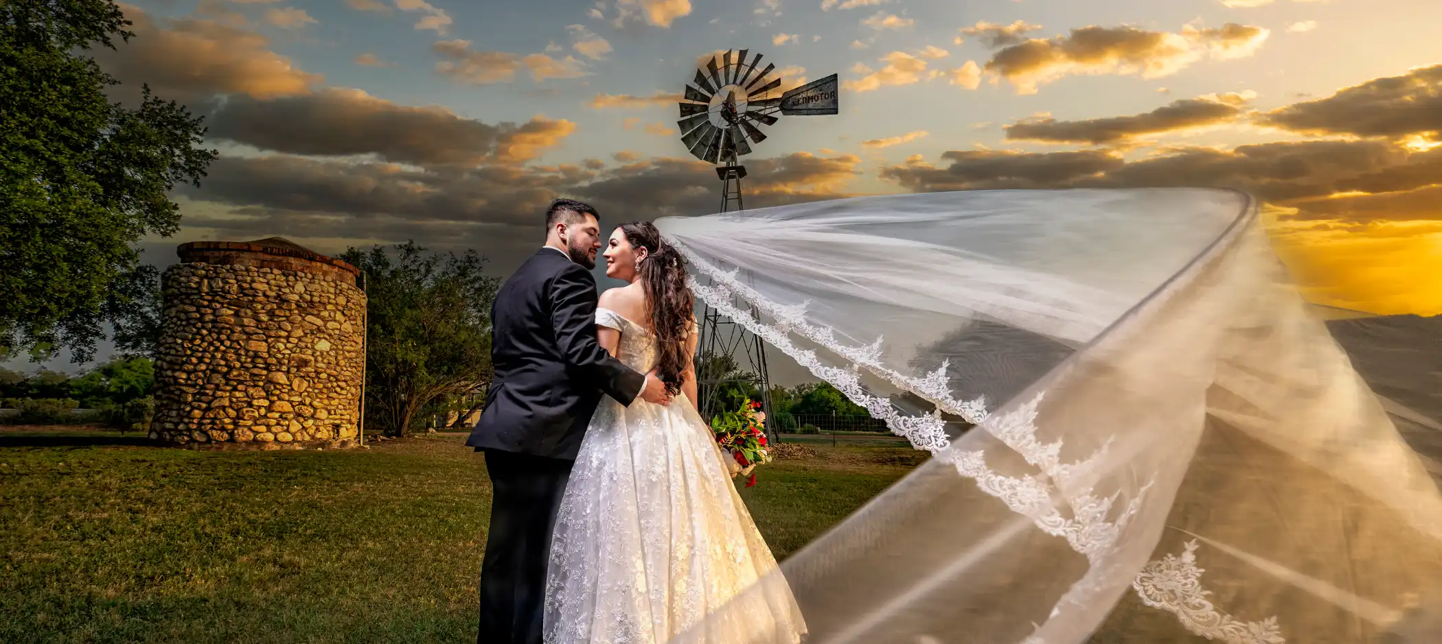 Professional Photographer A bride and groom embrace outdoors at sunset, their long veil flowing in the wind. A photographer captures this romantic moment near a windmill and stone building under a partly cloudy sky—beautiful memories in portraits. www.maranonimagery.com