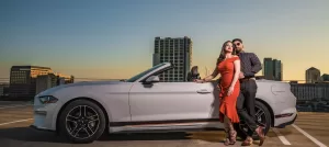 Professional Photographer A couple poses beside a white convertible car on a rooftop at sunset, with city buildings in the background. The woman in a red dress and the man in a dark shirt and sunglasses create striking portrait photography moments. www.maranonimagery.com