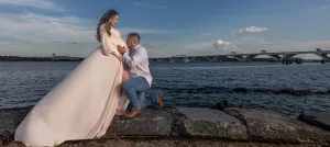 Professional Photographer A man kneels beside a pregnant woman in a flowing pale dress by the water, holding her belly. With a bridge and blue sky with clouds in the background, this scene beautifully captures the essence of wedding photography. www.maranonimagery.com