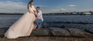 Professional Photographer A man kneels and kisses the belly of a pregnant woman in a flowing dress by the water, with a bridge and blue sky in the background—capturing a tender moment perfect for portraits. www.maranonimagery.com