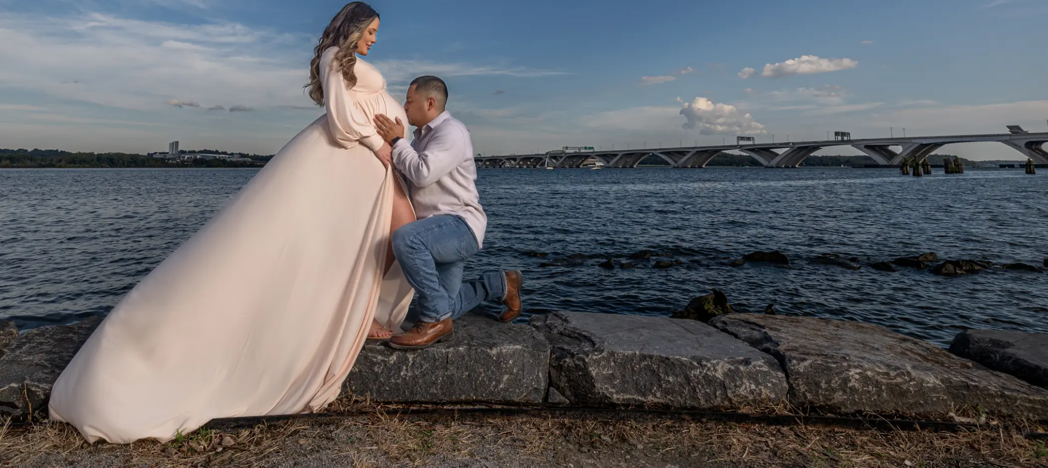 Professional Photographer A man kneels and kisses the belly of a pregnant woman in a flowing dress by the water, with a bridge and blue sky in the background—capturing a tender moment perfect for portraits. www.maranonimagery.com