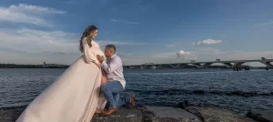Professional Photographer A man kneels on rocks beside a body of water, gently holding a pregnant woman's belly. Captured by a photographer, the woman wears a flowing light pink dress, with a large bridge stretching across the background under a partly cloudy sky. www.maranonimagery.com