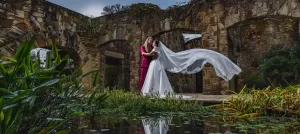 Professional Photographer A couple embraces outdoors by a stone ruin in this stunning wedding photography scene. One wears a white wedding dress with a long veil blowing in the wind; the other dons a magenta suit. Green plants and pond water complete the romantic setting. www.maranonimagery.com