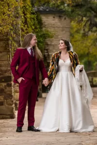 Professional Photographer A bride in a white dress and colorful fur shawl holds hands with a groom in a burgundy suit as they smile at each other outdoors near stone walls and greenery, creating timeless portraits full of love and joy. www.maranonimagery.com