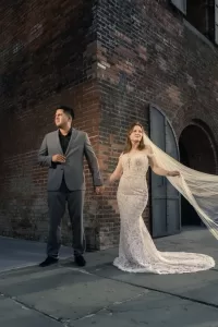 Professional Photographer A bride in a lace gown and long veil holds hands with a groom in a gray suit. They stand on a sidewalk by a brick building, gazing in different directions, softly illuminated—an elegant moment of wedding photography. www.maranonimagery.com