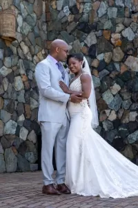 Professional Photographer A bride and groom smile and embrace on their wedding day, standing in front of a stone wall. Captured by a talented wedding photographer, the bride wears a white gown with a train and veil; the groom wears a light gray suit with a purple tie. www.maranonimagery.com