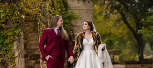 Professional Photographer A bride in a white dress and colorful shawl walks hand in hand with a groom in a burgundy suit, both smiling at each other as their wedding photographer captures the moment in a lush, outdoor setting with stone walls and greenery. www.maranonimagery.com