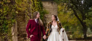 Professional Photographer A bride in a white dress and patterned shawl holds her gown while walking hand-in-hand with a groom in a burgundy suit outdoors near a stone wall covered in greenery. Both are smiling at each other, perfectly captured by skilled wedding photography. www.maranonimagery.com
