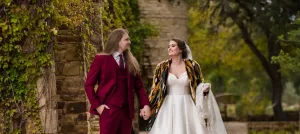 Professional Photographer A bride in a white gown and colorful shawl holds hands with a groom in a burgundy suit as they walk outdoors near a stone building surrounded by greenery, both smiling at each other—capturing timeless wedding photography moments. www.maranonimagery.com