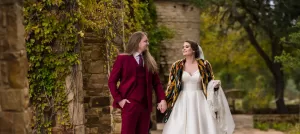 Professional Photographer A bride in a white dress and colorful shawl smiles while holding hands with a groom in a burgundy suit as they walk outdoors by a stone wall covered in green vines—a stunning moment of wedding photography. www.maranonimagery.com