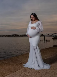 Professional Photographer A photographer captures a pregnant woman in a long, white fitted gown by the waterfront at sunset, gently holding her belly and smiling, with a bridge and calm water in the background. www.maranonimagery.com