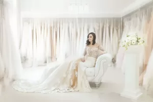 Professional Photographer A bride in a long, elegant lace wedding gown sits on a white upholstered chair in a bright room filled with hanging wedding dresses—a scene perfect for stunning wedding photography. A pedestal with a floral arrangement is beside her. www.maranonimagery.com