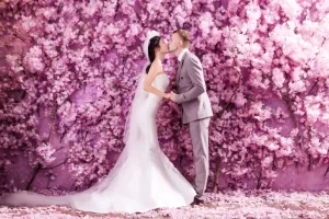 Professional Photographer A bride and groom share a kiss in front of a vibrant wall covered with pink cherry blossoms. Capturing this tender moment, the wedding photography highlights her flowing white gown and veil, and his light gray suit. www.maranonimagery.com