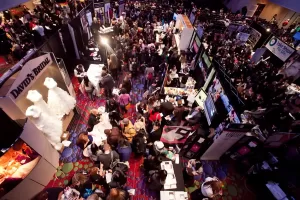 Professional Photographer A crowded convention or expo hall seen from above, with many people visiting various booths and displays, including one featuring portraits and headshots—and a stunning white wedding dress on the left side. www.maranonimagery.com
