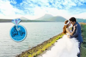 Professional Photographer A bride and groom embrace by a serene lakeside with mountains in the background. The image features a “She Said Yes Wedding Expo” logo shaped like a diamond ring on the left side, showcasing expert portraits for your special day. www.maranonimagery.com