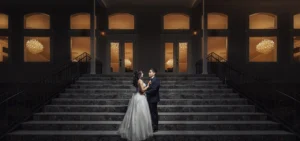 Professional Photographer A bride and groom stand on a grand stone staircase at night, facing each other and holding hands; behind them, large windows glow warmly, revealing bright chandeliers inside—a timeless moment of wedding photography. www.maranonimagery.com