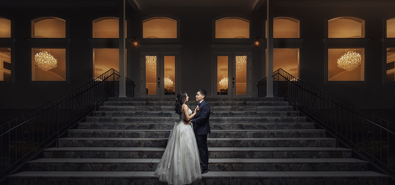 Professional Photographer A bride and groom stand on a grand stone staircase at night, facing each other and holding hands; behind them, large windows glow warmly, revealing bright chandeliers inside—a timeless moment of wedding photography. www.maranonimagery.com