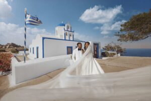 Professional Photographer A bride in a white wedding gown and a groom in a white tuxedo stand together in front of a white and blue chapel in Greece, with the brides long veil trailing across the foreground. www.maranonimagery.com